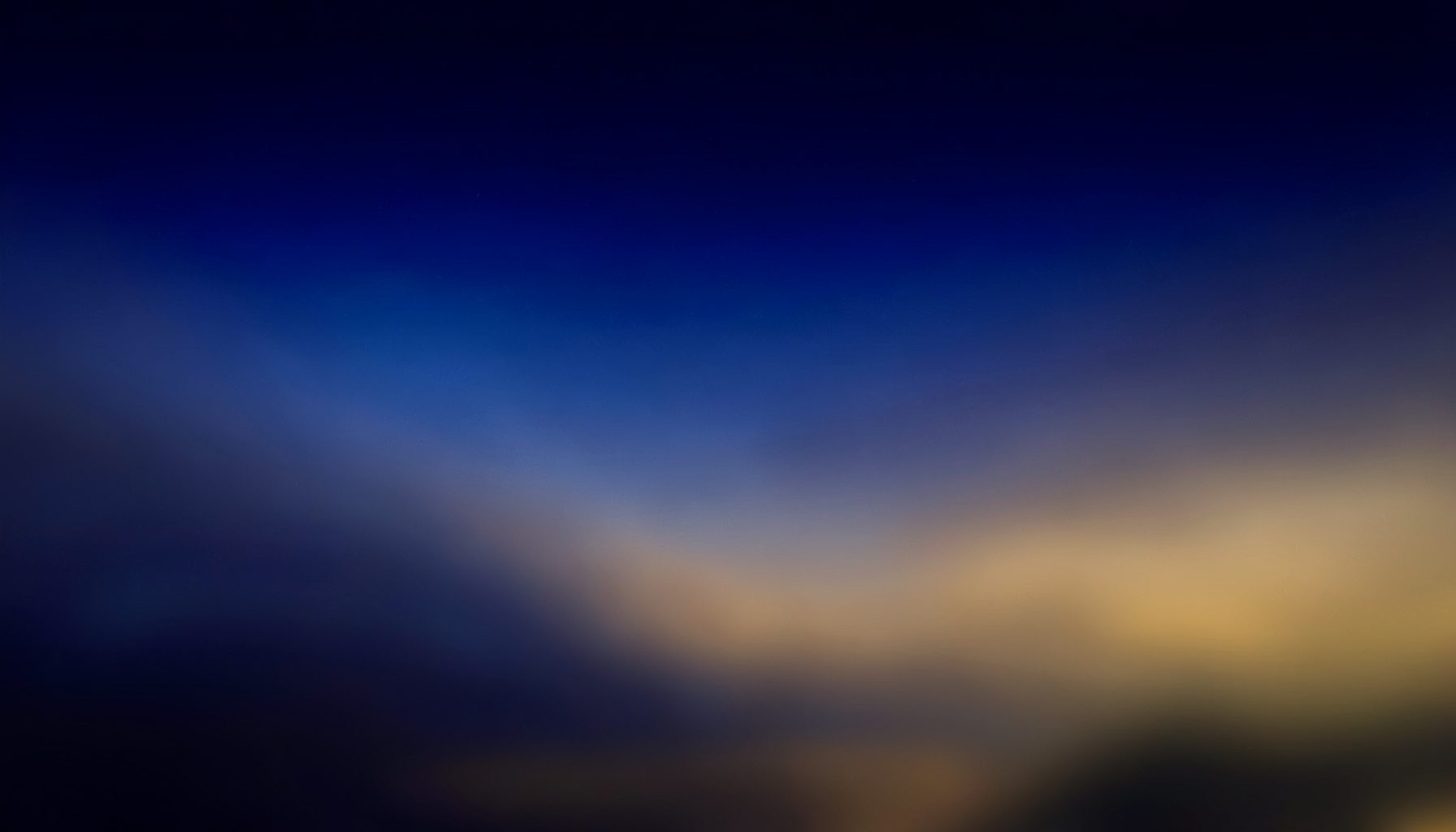 A wide atmospheric composition of deep indigo and navy tones blending into muted amber and gold at the horizon, suggesting festival lights against a night sky, with soft haze and no figures.